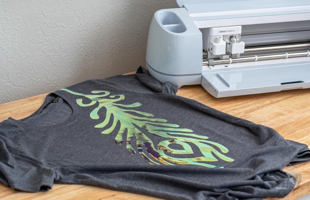 Cricut Maker 3 on table next to  gray shirt with holographic feather on it. 