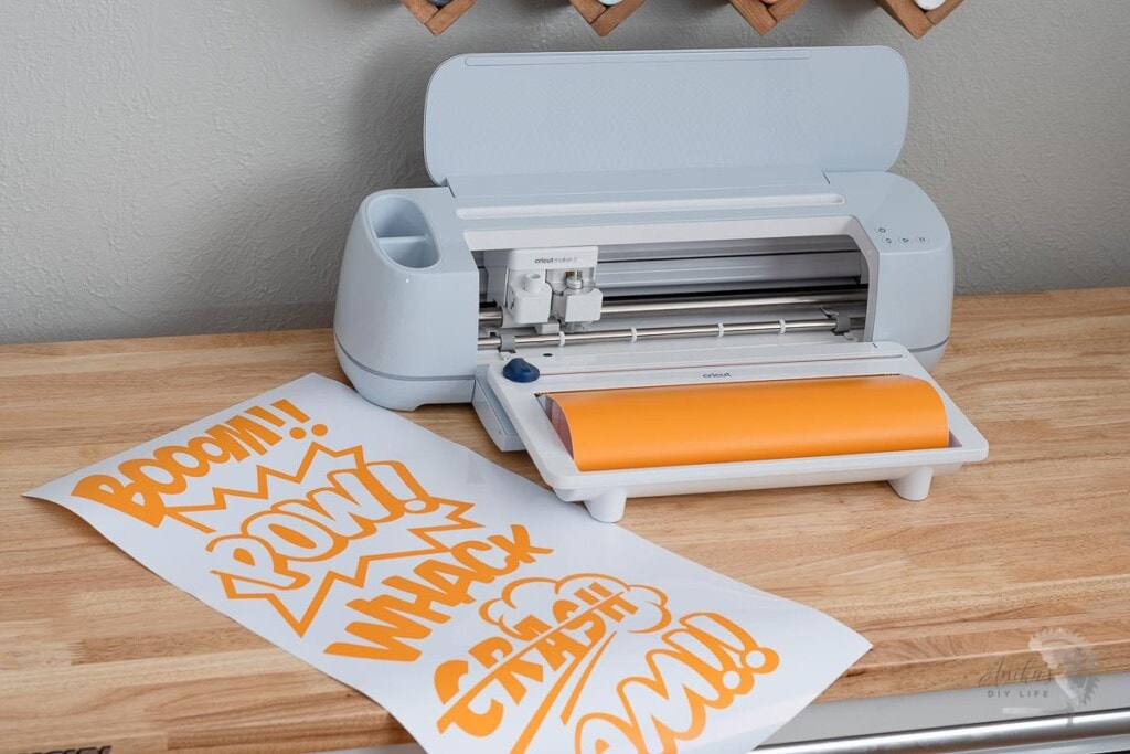 Cricut Maker 3 on Table with orangle roll and large comic book word art cut out next to it